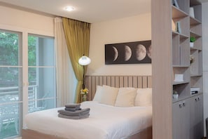 A bedroom with a bed, a nightstand, a wall-mounted shelf, and a window with curtains. - Spacious Studio with Balcony in Nimman Soi 15 | Pool, Gym & Washer (Chiang Mai)