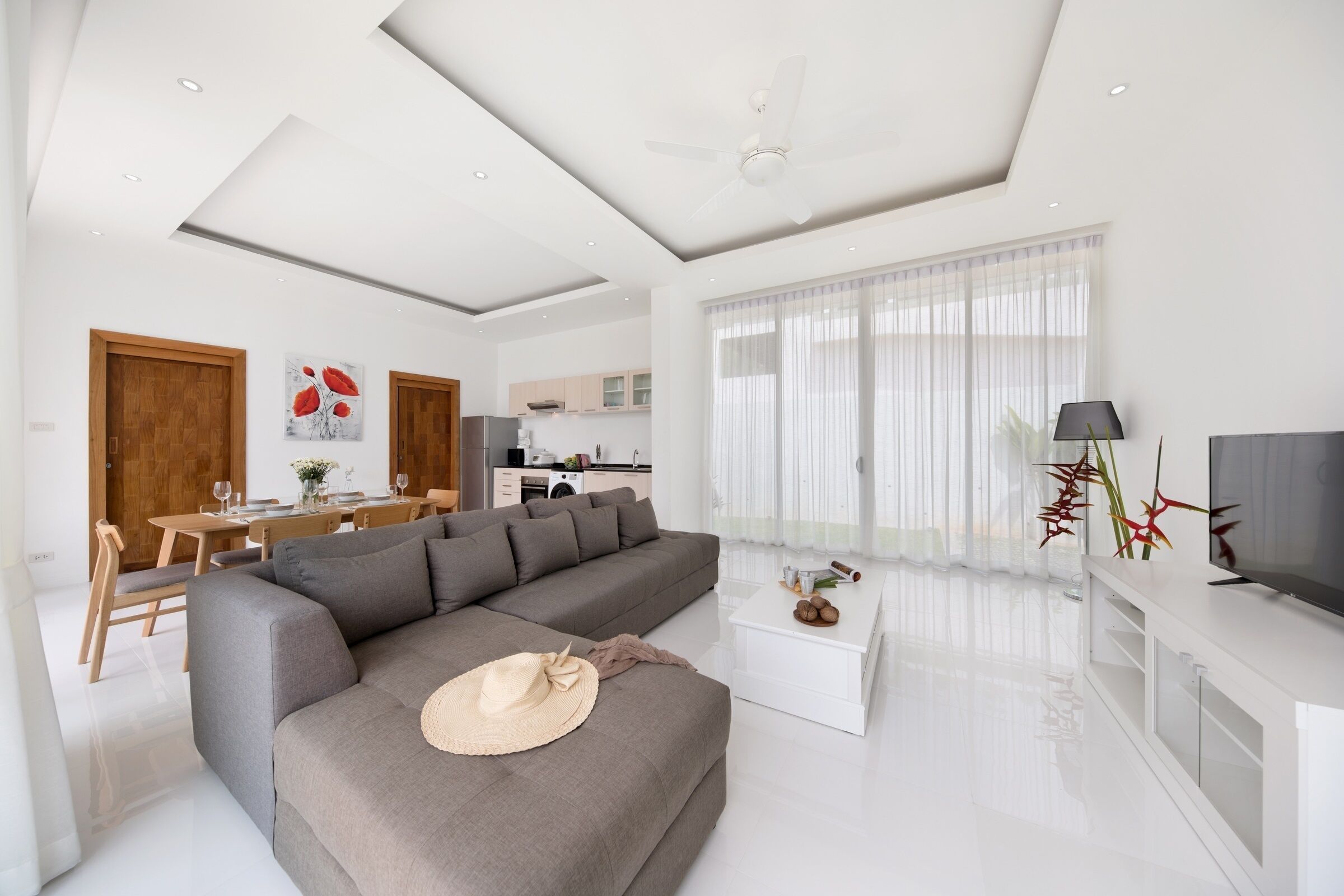 A modern living room with a grey sofa, a dining area with wooden furniture, a kitchen in the background, and a TV on a white stand.