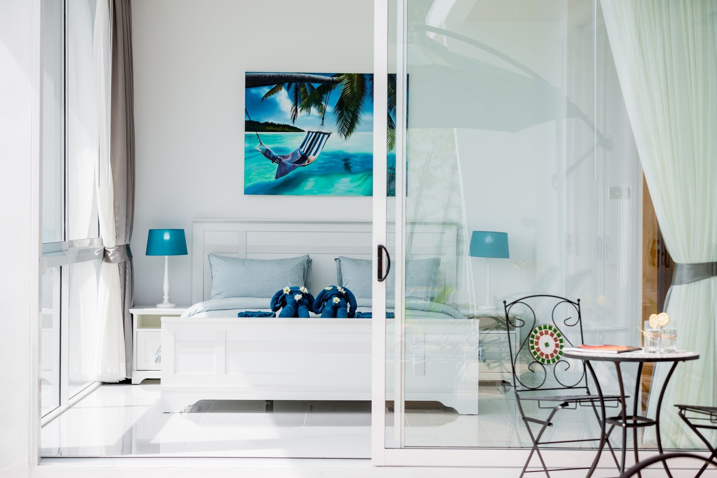 A bedroom with a bed, two bedside lamps, a glass door leading to a balcony, and a painting of a beach scene.