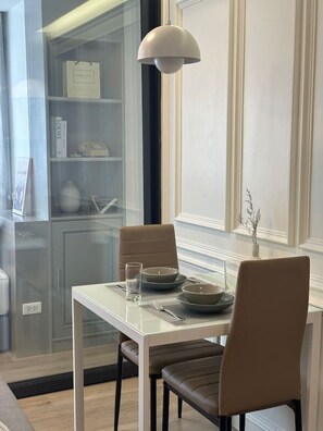 Ein Essbereich mit einem Glastisch, zwei Stühlen, einer Pendelleuchte und Blick auf eine Küche mit Schränken und einem Kühlschrank.