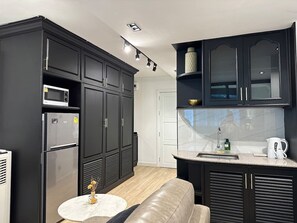 Una cocina moderna con armarios oscuros, microondas integrado, refrigerador de acero inoxidable, una mesa redonda blanca con un adorno y una encimera con hervidor y dispensador de jabón.
