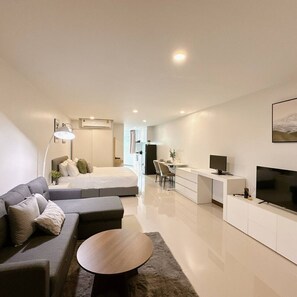 A modern, well-lit living room with a sofa, a round coffee table, a bed, a desk with a chair, and a TV. - Elegant Modern White Suite (Chang Phueak)