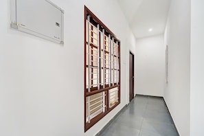 Un pasillo con un gran ventanal de madera y persianas blancas, una puerta al final y un interruptor de luz en la pared.