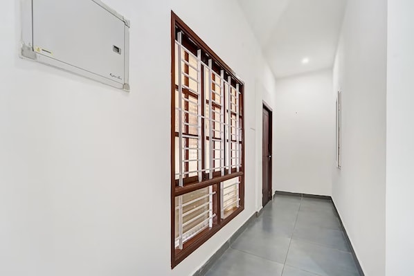 A hallway with a large window with a wooden frame and white blinds, a door at the end, and a light switch on the wall.