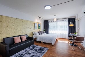 Una habitación moderna con pared de ladrillo, una cama, sofá negro, mesa de comedor con sillas y un ventanal con cortinas.