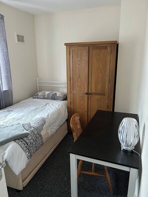 A bedroom with a bed, a wooden wardrobe, a table with a lamp, and a chair.