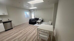 Een moderne keuken met een vaatwasser, een eethoek met een witte tafel en stoel, een zwarte bank en een raam met jaloezieën.