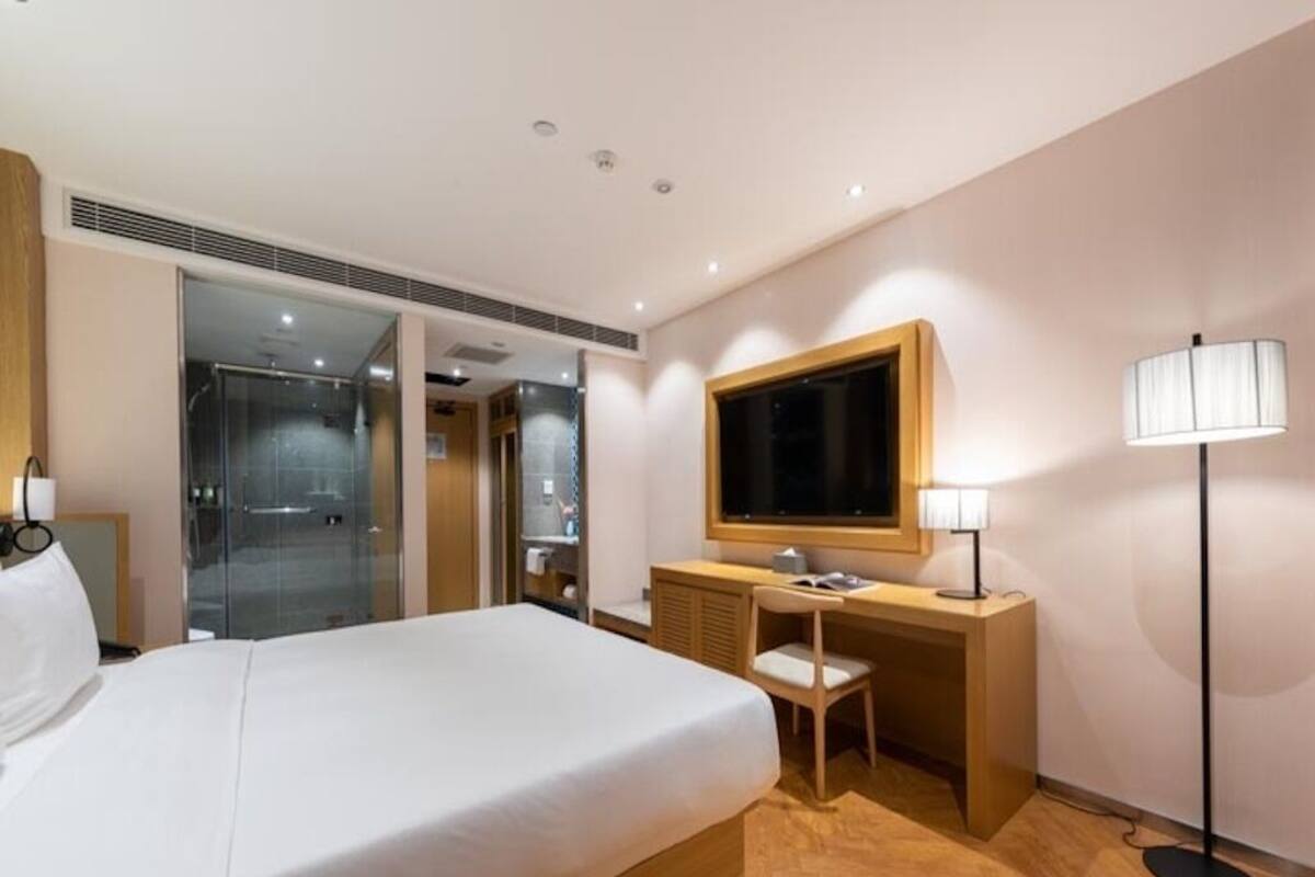 A hotel room with a bed, desk, chair, TV, lamp, and a glass shower area.