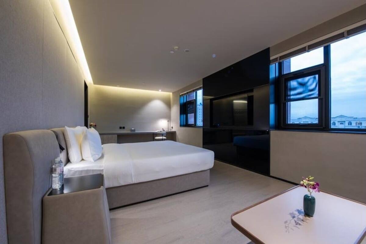 A modern hotel room with a bed, a seating area, a small table with a vase, a TV, and a window with a view.