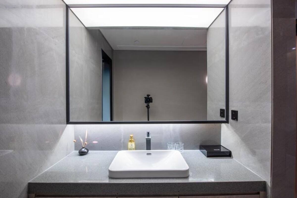 A modern bathroom with a large mirror, a white sink, a small vase, and a soap dispenser.