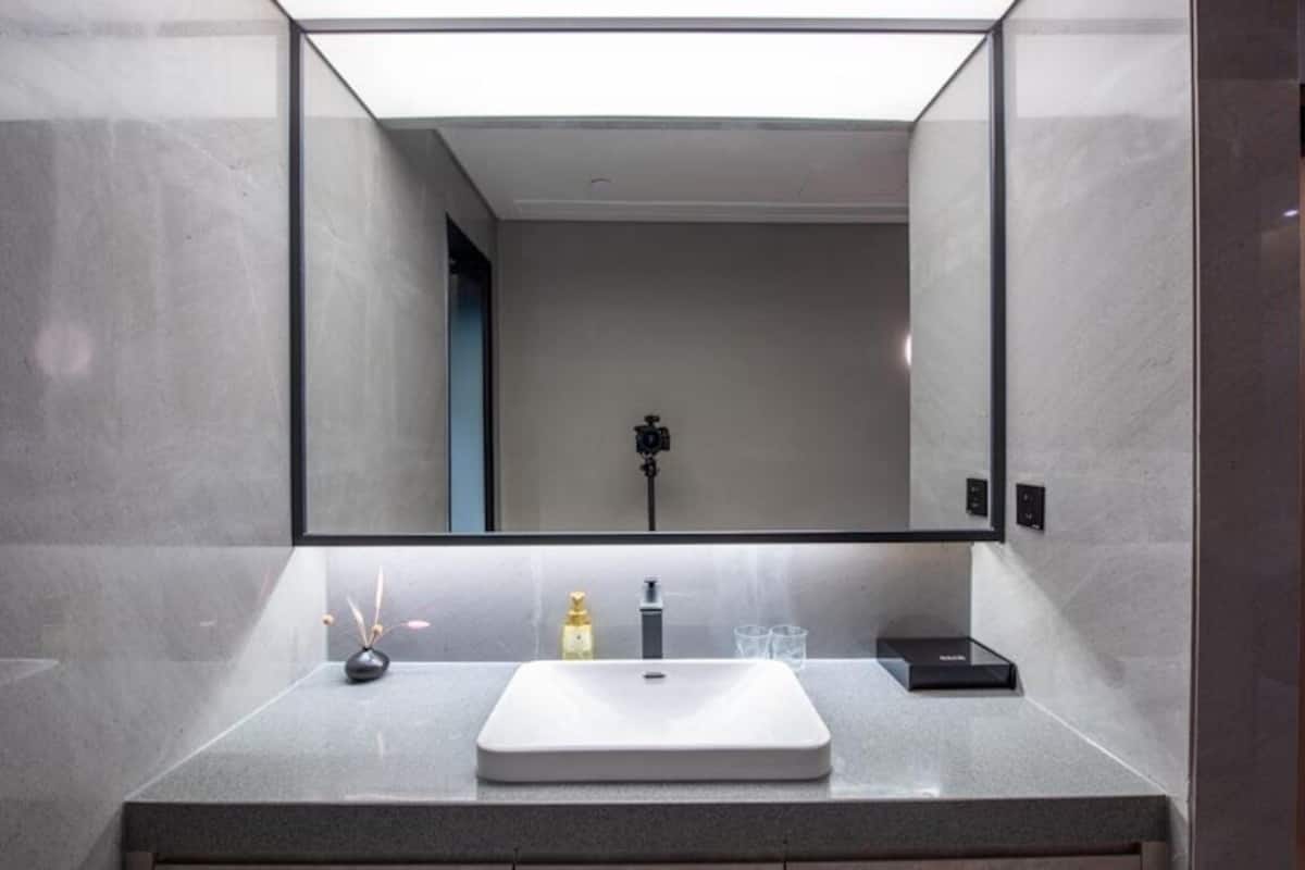 A modern bathroom with a large mirror, a white sink, a small vase, and a soap dispenser.