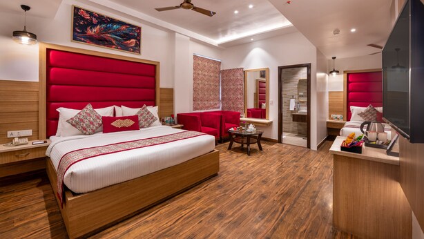 Quarto de hotel moderno com uma cama grande, cabeceira estofada em vermelho, ventilador de teto e um pequeno espaço de estar.