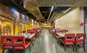 Restaurante com cadeiras e mesas estofadas em vermelho, área de bar central e murais decorativos nas paredes.