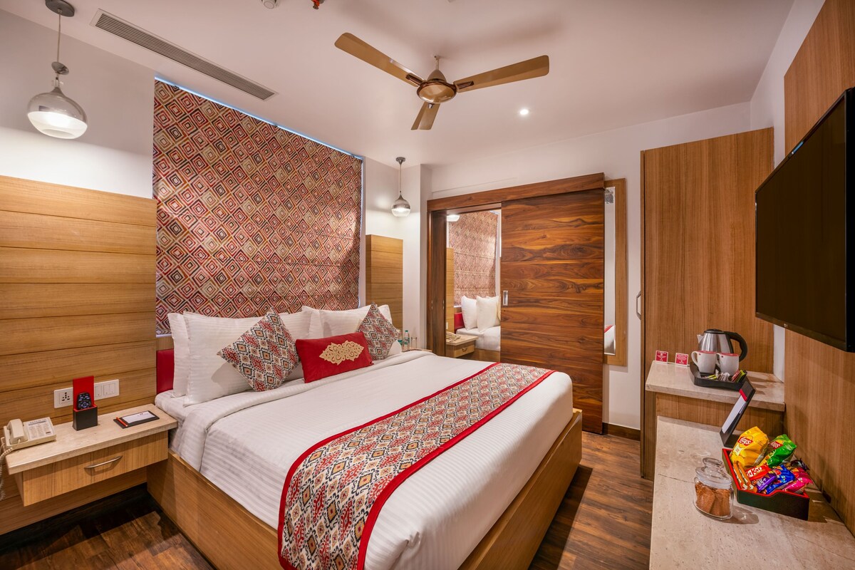 A modern hotel room with a large bed, a ceiling fan, a wooden headboard, a bedside table with a phone and a red box, and a small table with a kettle and snacks.