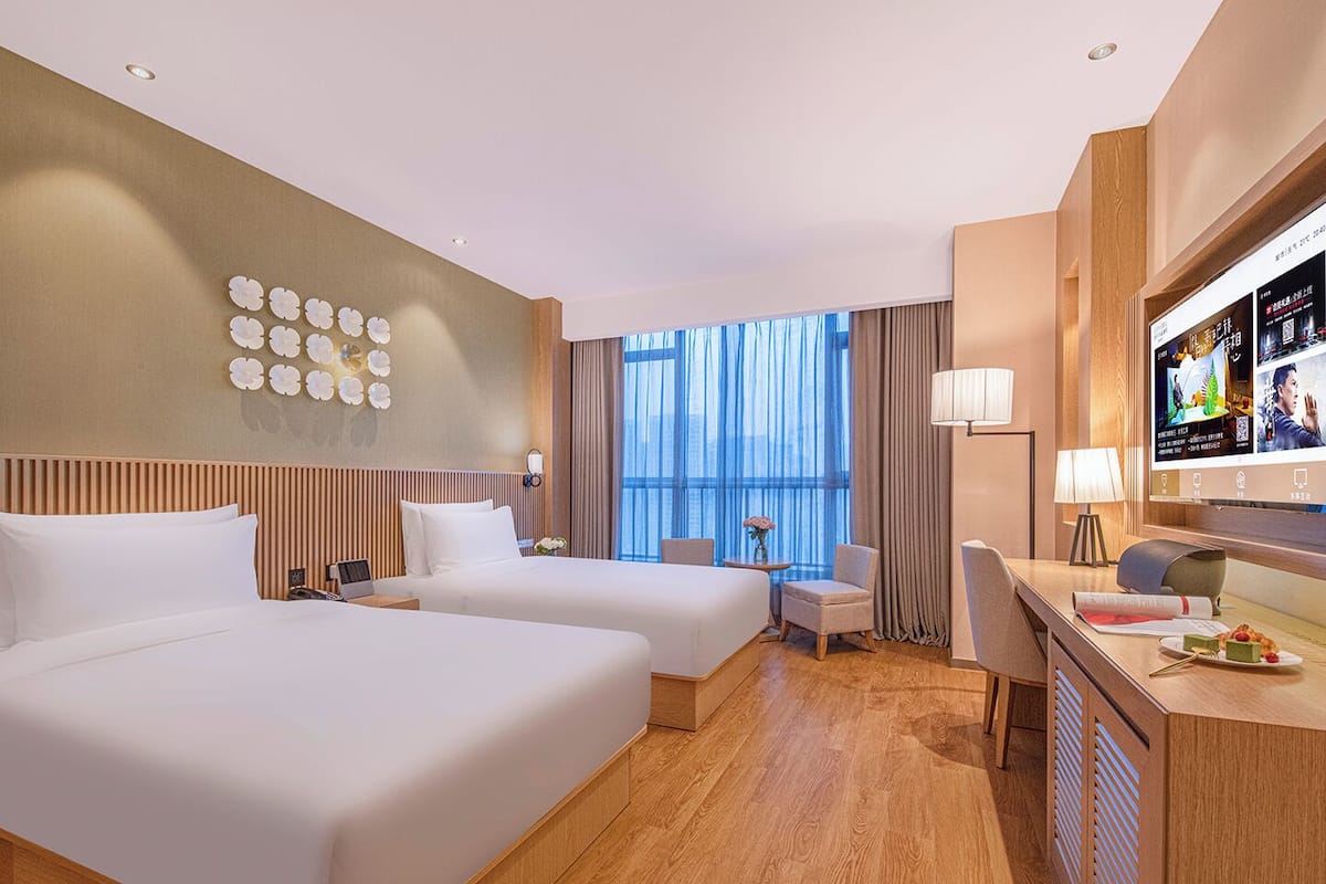 Habitación de hotel con dos camas, un escritorio con televisor, una silla, una mesita con flores y un ventanal con cortinas.
