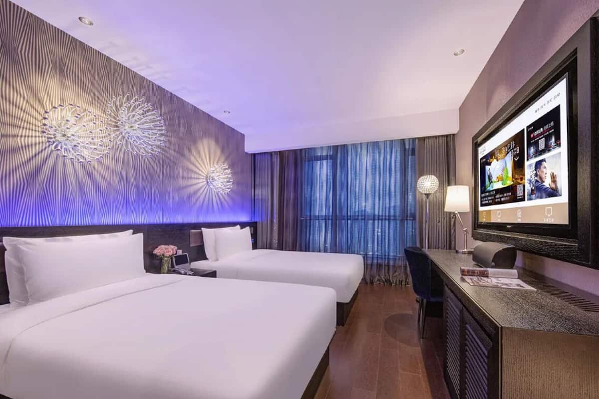 Habitación de hotel con dos camas, un escritorio, un televisor y una pared con diseño decorativo.