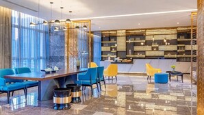A modern hotel lobby with a large dining table, blue and yellow chairs, a bar area, and a decorative wall with shelves.