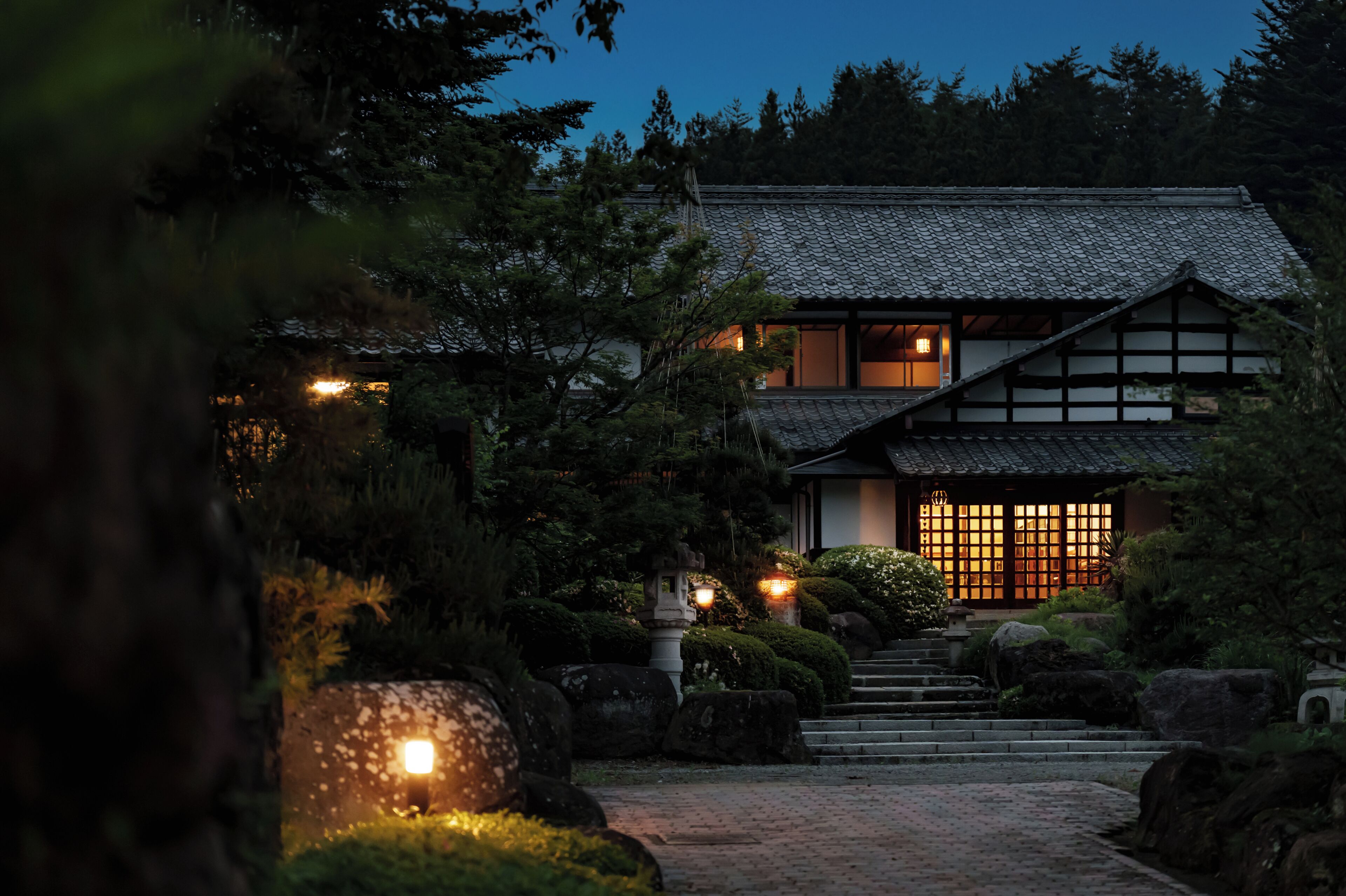 Ett traditionellt japanskt hus med ett tak täckt av tegelpannor, en träkonstruktion och en stensatt gångväg som leder till entrén.