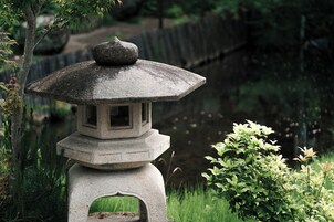 Un farol de piedra con diseño tradicional, ubicado en un jardín con cerca de madera y vegetación al fondo.