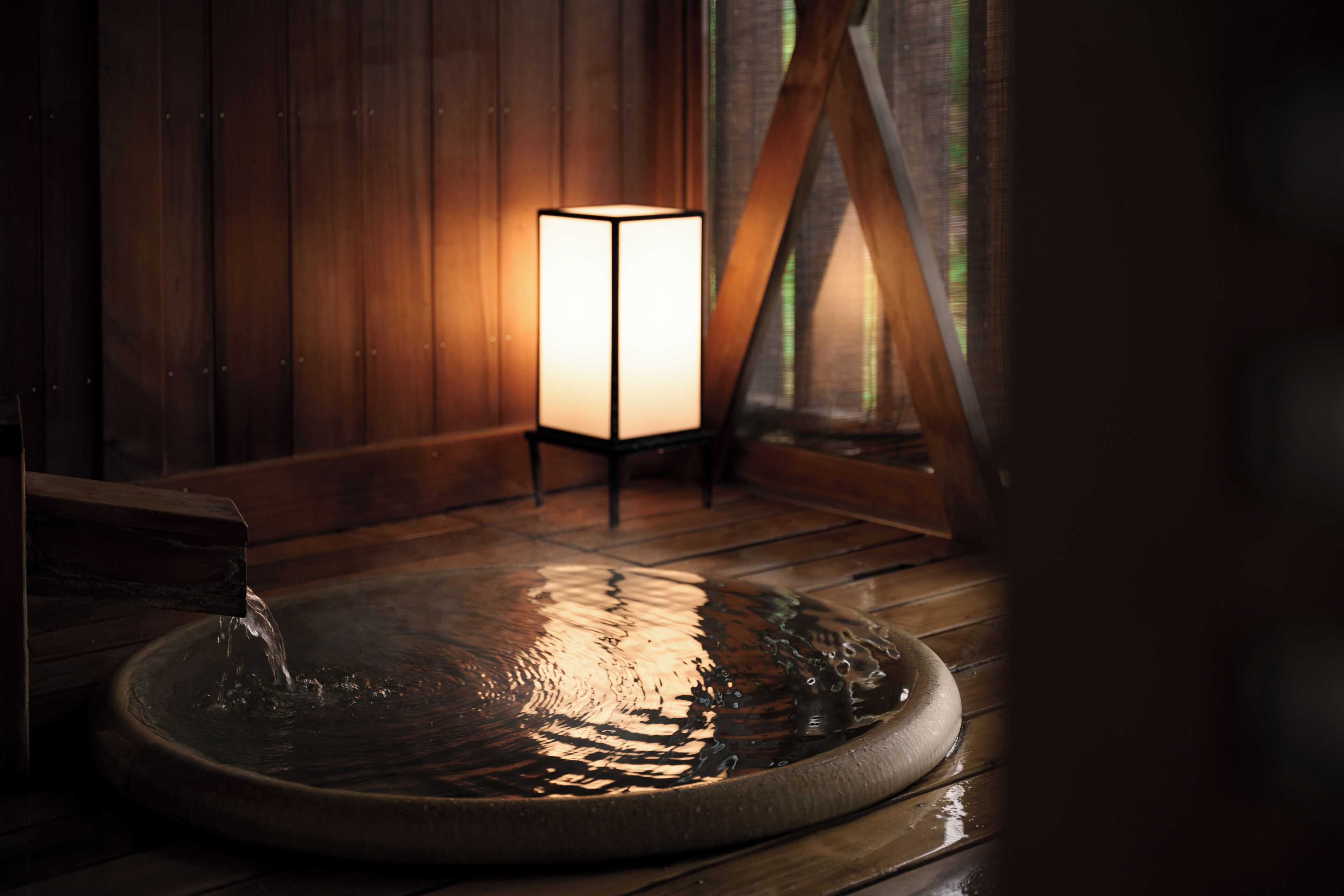 A wooden interior with a hot spring bath, a standing lamp, and a window with a view.
