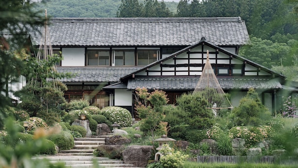 A traditional Japanese house with a tiled roof, surrounded by lush greenery and a stone staircase leading to the entrance.