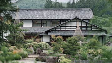 Une maison traditionnelle japonaise, avec un toit recouvert de tuiles, entourée d’une végétation luxuriante et dotée d’un escalier en pierre menant à l’entrée.