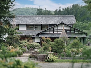 Ein traditionelles japanisches Haus mit einem Ziegeldach, umgeben von üppiger Vegetation und einer Steintreppe, die zum Eingang führt.