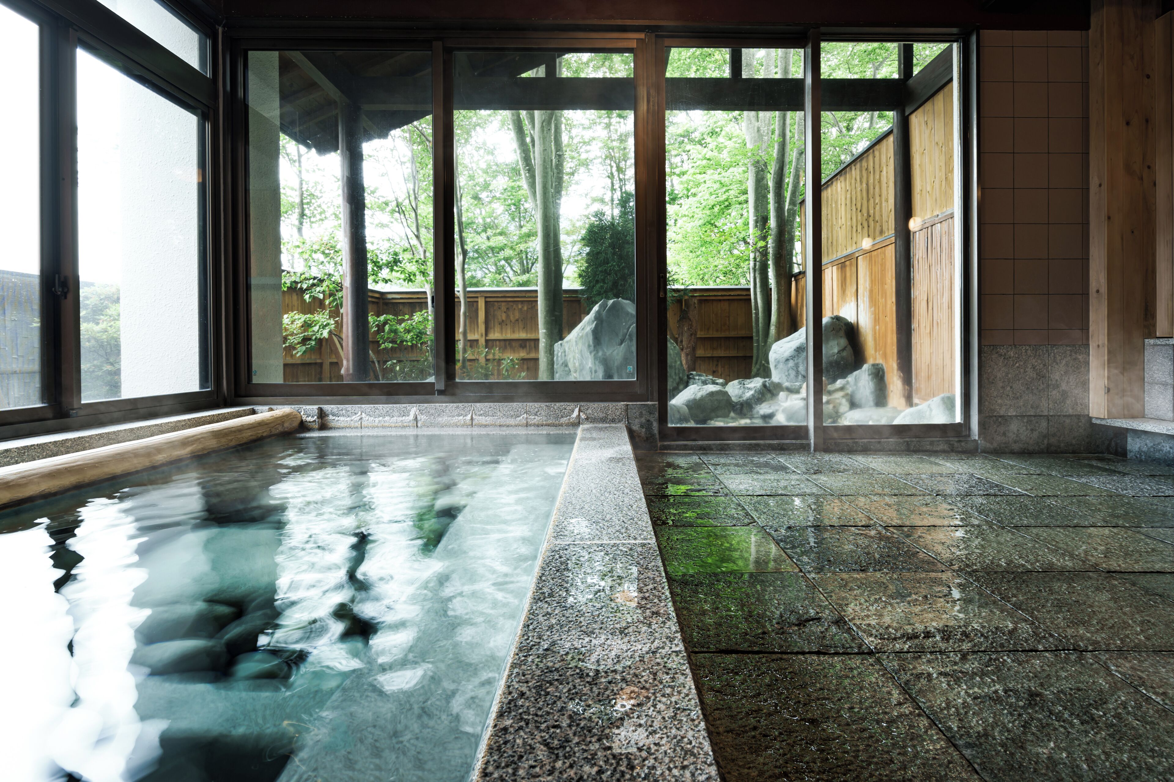 A traditional Japanese onsen with a stone bath, wooden deck, and a view of a forested area through large sliding doors.