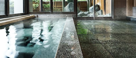 A traditional Japanese onsen with a stone bath, wooden deck, and a view of a forested area through large sliding doors.