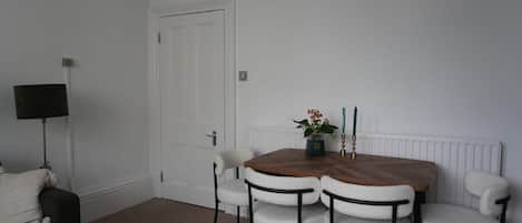Una habitación con una mesa de comedor, cuatro sillas, una lámpara de pie negra, una puerta blanca y un radiador.