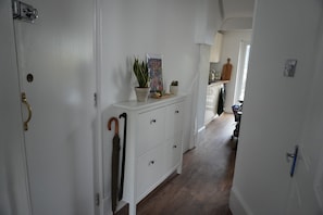 Een gang met een witte deur, een wit kastje met lades, een potplant en een wandelstok die tegen de muur staat.