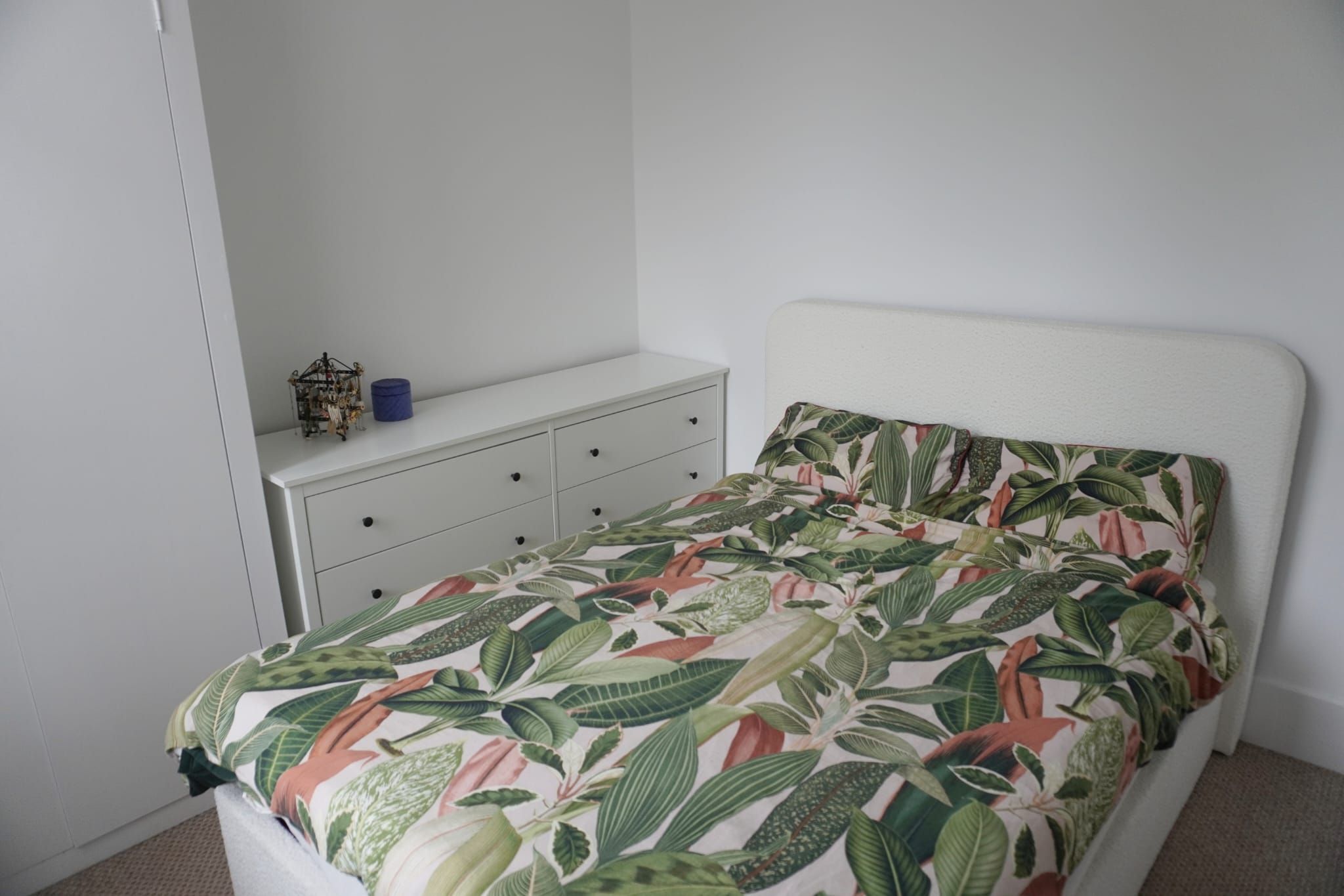 A bedroom with a bed, a white dresser, and a small shelf with a blue container.