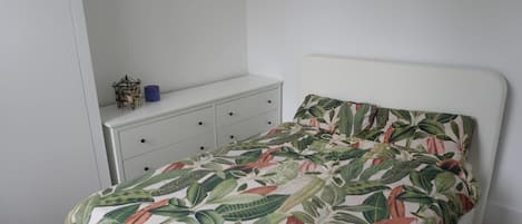 A bedroom with a bed, a white dresser, and a small shelf with a blue container.