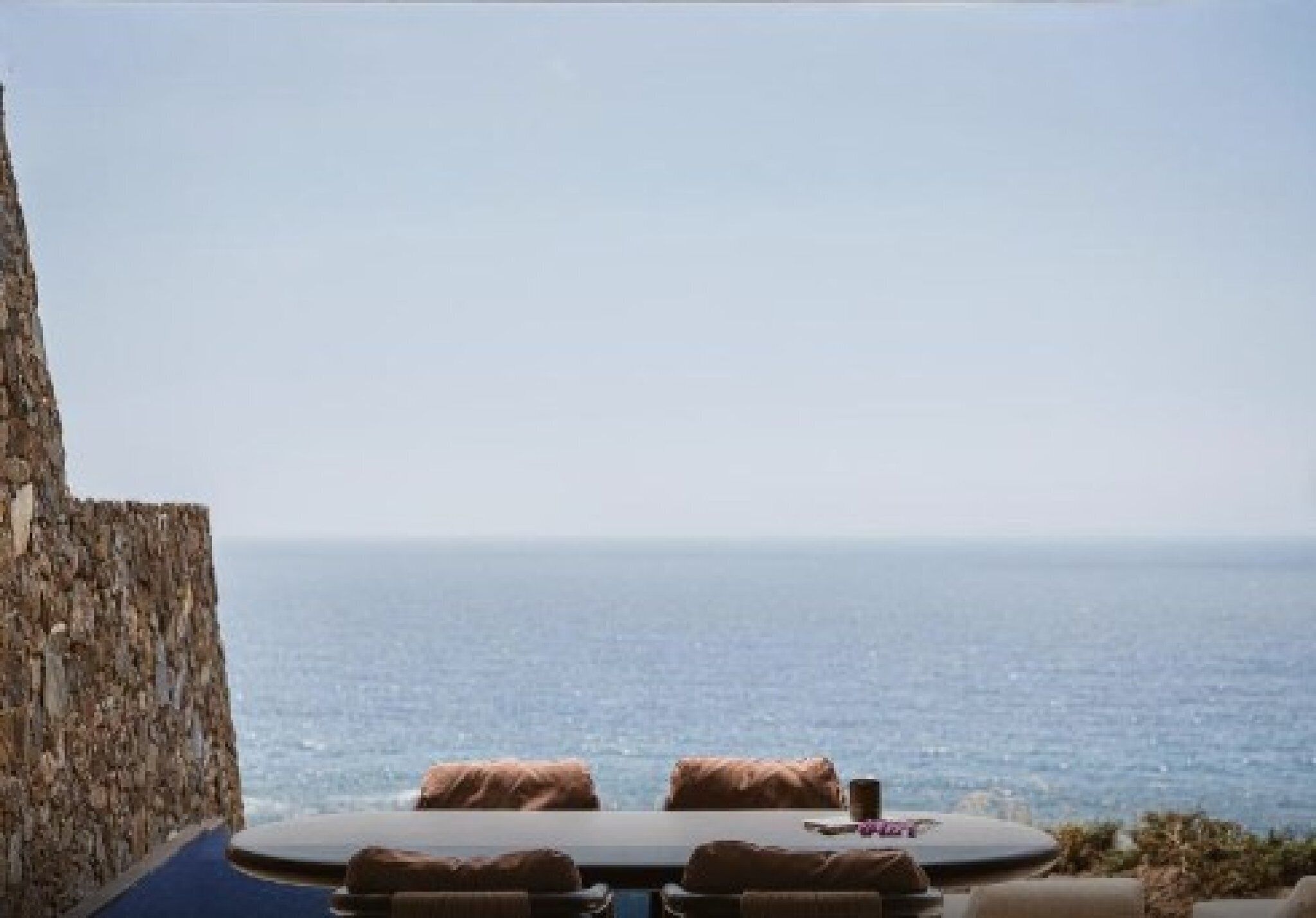 A stone wall, a round table with cushions, and a view of the sea.