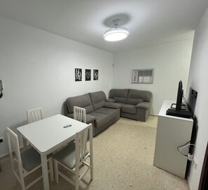 A small, sparsely furnished room with a white table, two chairs, a gray sofa, a small refrigerator, and three wall-mounted pictures.