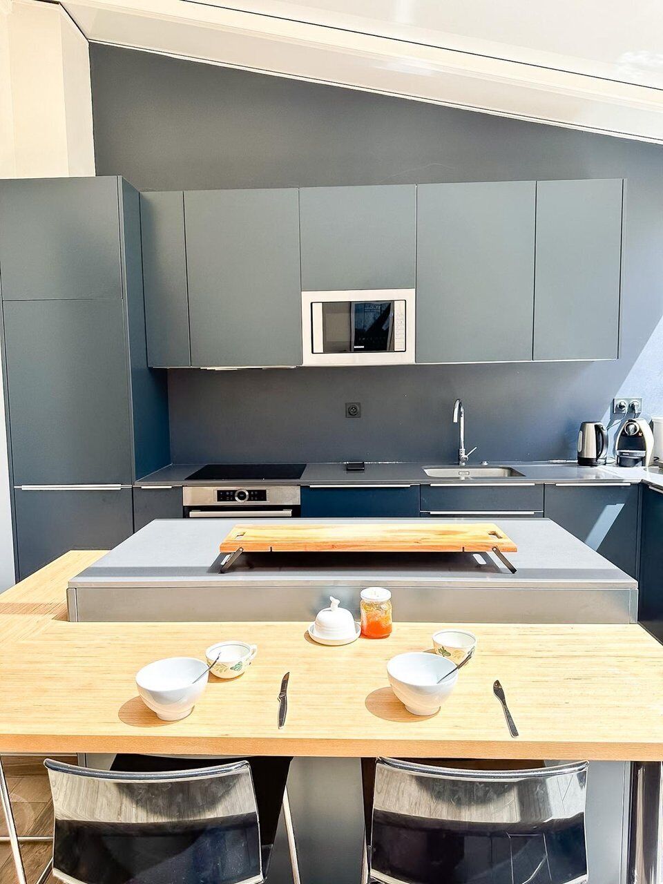 A modern kitchen with grey cabinets, a microwave, a sink, and a wooden table with bowls and utensils.