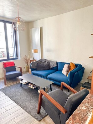 Een woonkamer met een blauwe bank, een salontafel en een stoel. Er staat een lamp op een bijzettafeltje, een raam met jaloezieën en een radiator.