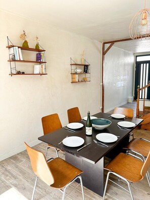 Een eethoek met een lange tafel gedekt voor zes personen, omringd door houten stoelen. Twee planken aan de muur met boeken en decoratieve objecten.