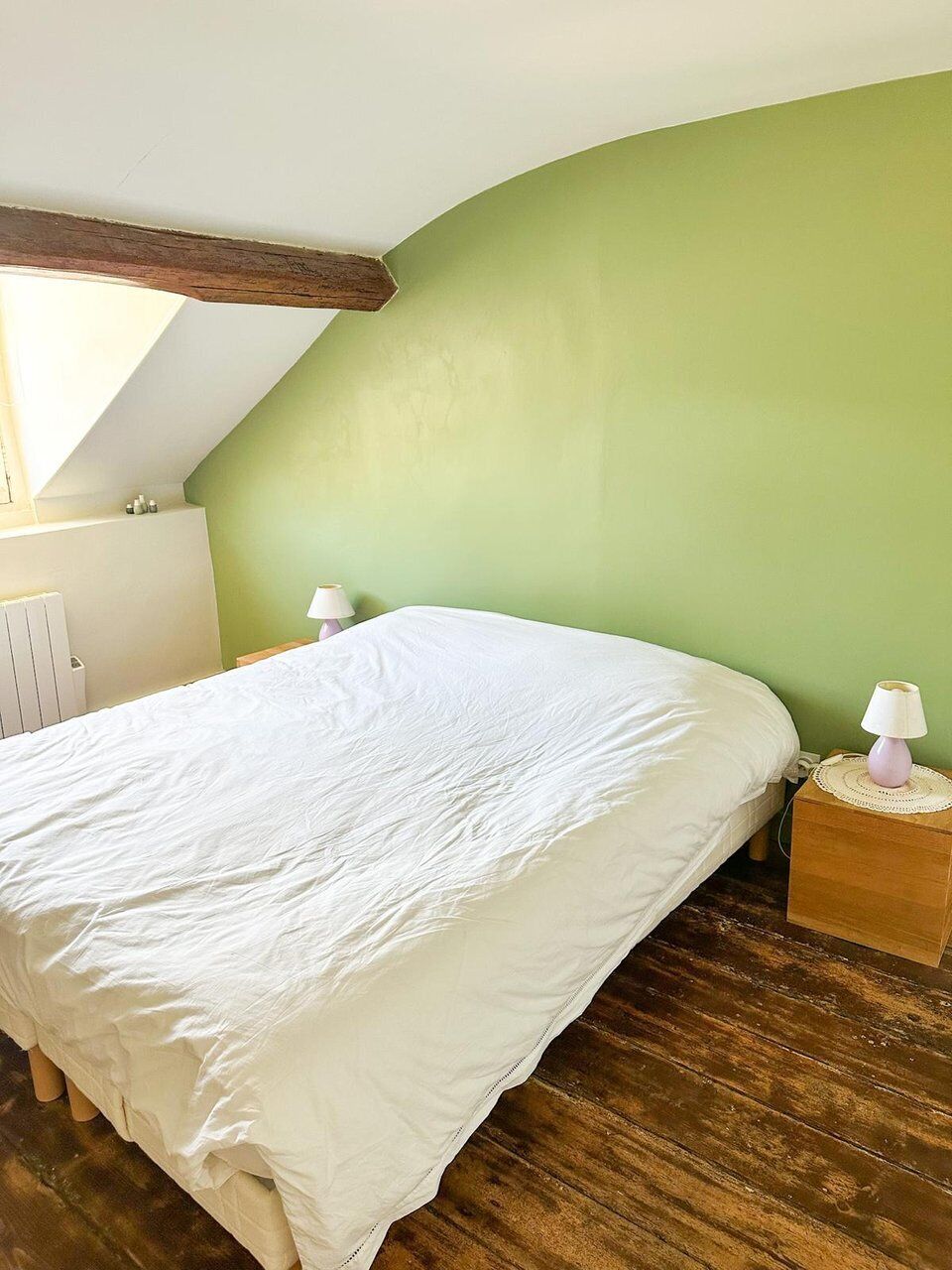 A bedroom with a sloped ceiling, a bed with white bedding, a nightstand with a lamp, and a radiator.