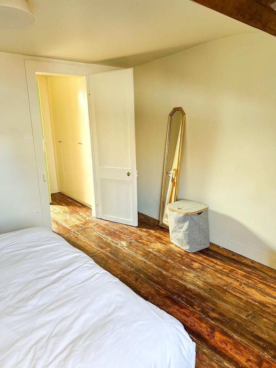 A bedroom with a wooden floor, a bed, a door, and a mirror.