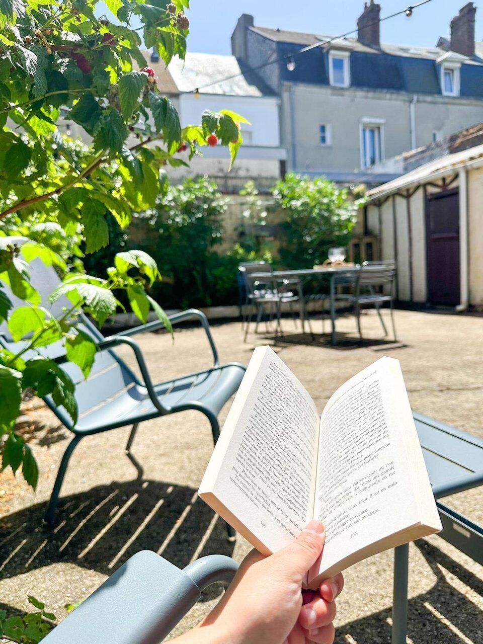 A person reading a book outdoors with a table and chairs in the background.
