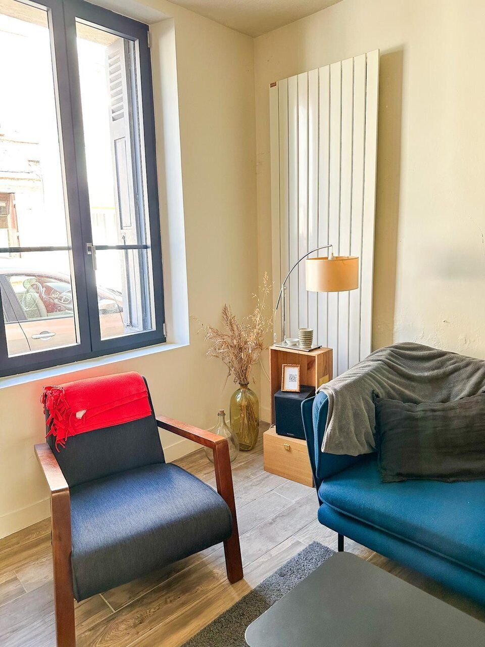 Une pièce avec une fenêtre, un canapé bleu, un fauteuil rouge, une lampe et un radiateur.