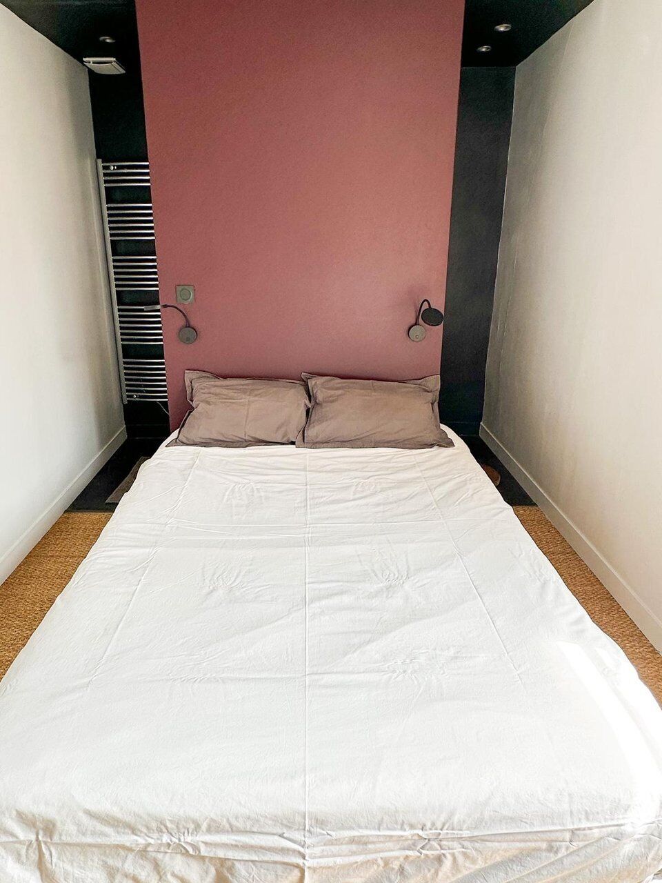 Ein schmales Zimmer mit einem Bett, einer rosa Wand und einer schwarzen Säule.