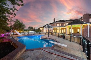 Une piscine avec un toboggan, entourée d’une terrasse en briques, et un bâtiment à la façade en briques et aux éclairages décoratifs.