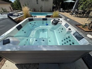 Ein Whirlpool mit Bedienfeld, umgeben von einer Terrasse mit Gartenmöbeln und einem Pool.