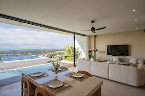 Una sala moderna con un televisor grande, un comedor con una mesa preparada para dos personas, y vistas a una piscina y paneles solares.