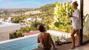 Ein Mann sitzt am Pool mit Blick auf eine Stadtlandschaft, eine Frau steht mit einem Getränk in der Hand, und im Hintergrund steht eine tropische Pflanze.