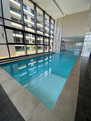 Ein Schwimmbad mit Fliesenboden, großen Fenstern und Blick auf ein mehrgeschossiges Gebäude.