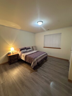 A bedroom with a bed, a nightstand with a lamp, a window with blinds, and a ceiling light.