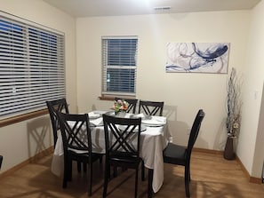 Un coin repas avec une table dressée pour quatre personnes, entourée de chaises, une fenêtre avec des stores et une œuvre d’art abstraite accrochée au mur.